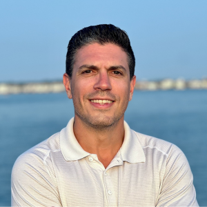 A person in a white polo shirt stands smiling with arms crossed in front of a calm body of water on a clear day.